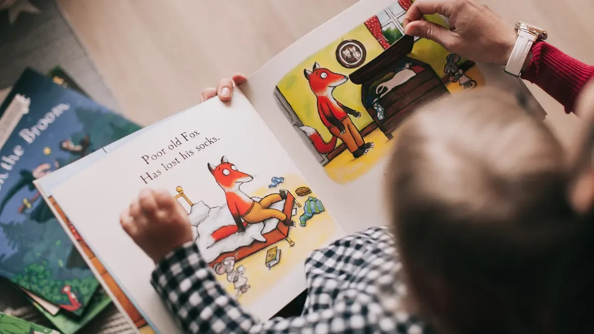 Criança sorrindo enquanto lê um livro com um adulto, demonstrando o prazer da leitura no processo de alfabetização e letramento.